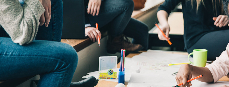 Four people are sitting at a desk writing something on a poster. There are also pencils and a cup on the table.